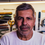 A confident middle-aged man with greying hair and a beard, wearing a white t-shirt, standing in a workshop setting, symbolizing the experienced and trustworthy professionals at ohmycleaners.com providing chimney cleaning services in Omaha